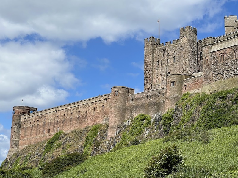 Bamburgh Castle stands majestically atop a green hill under a partly cloudy sky. The fortress features tall stone walls, towers, and battlements, showcasing its historical architecture.