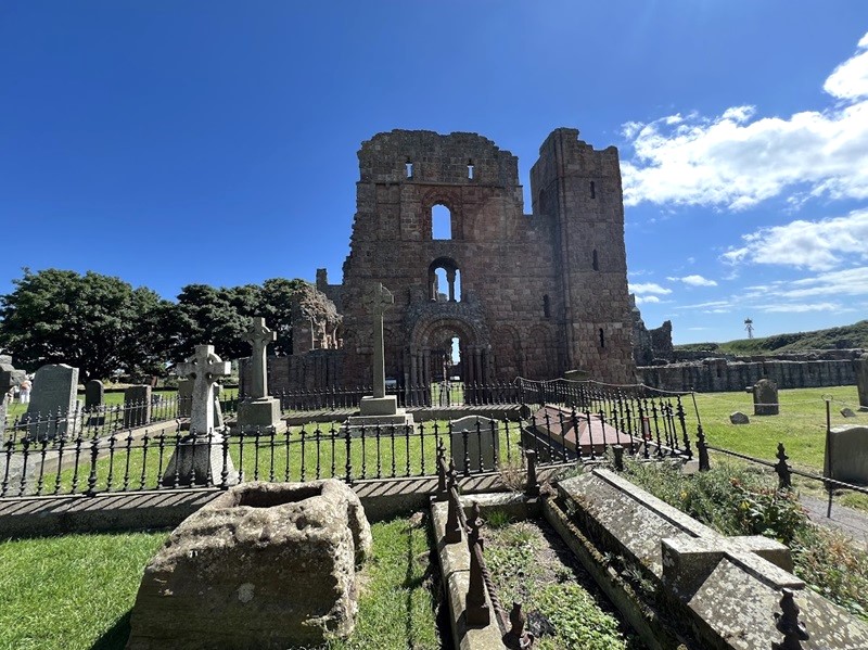 The ruins of an ancient stone building The ruins of Lindisfarne Priory on Holy Island, stand tall under a bright blue sky. Surrounding the structure is a graveyard with several tombstones and iron railings. The remnants of the building showcase archways and window openings, suggesting its historical significance.