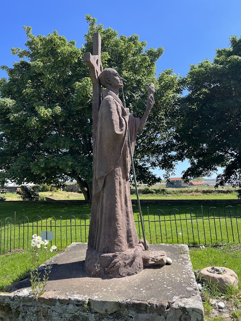 A depiction of St. Aidan standing on Holy Island, also known as Lindisfarne. He is shown in traditional monastic robes, holding a staff and gazing out towards the sea. 