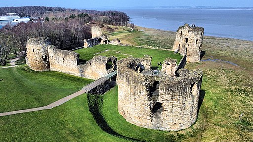 Aerial view of Flint Castle ruins, featuring large circular stone towers and walls, surrounded by green grass and located near a coastal shoreline. The castle has an open layout with visible remnants of the fortification, and a path leading into the ruins. The surrounding landscape includes trees and water in the background.