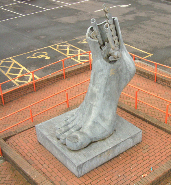 A large outdoor sculpture depicting a human foot with mechanical components emerging from the top. The foot appears severed at the ankle, and gears, pipes, and chains are visible protruding from the top of the sculpture. It is placed on a raised brick platform surrounded by orange railings, and the background shows an empty parking lot with yellow accessibility markings.