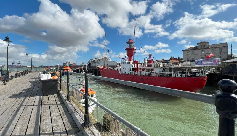 A bright red lightship moored at a pier in Harwich, UK, on a sunny day with blue skies and scattered clouds. The ship is docked alongside a wooden promenade with benches, railing, and street lamps.