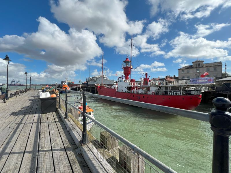 A bright red lightship moored at a pier in Harwich, UK, on a sunny day with blue skies and scattered clouds. The ship is docked alongside a wooden promenade with benches, railing, and street lamps.