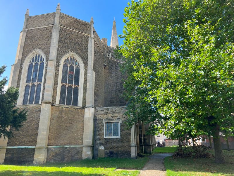 The exterior of St. Nicholas Church featuring large gothic-style windows, brick walls, and a tall spire. A tree provides shade in the churchyard under a clear blue s