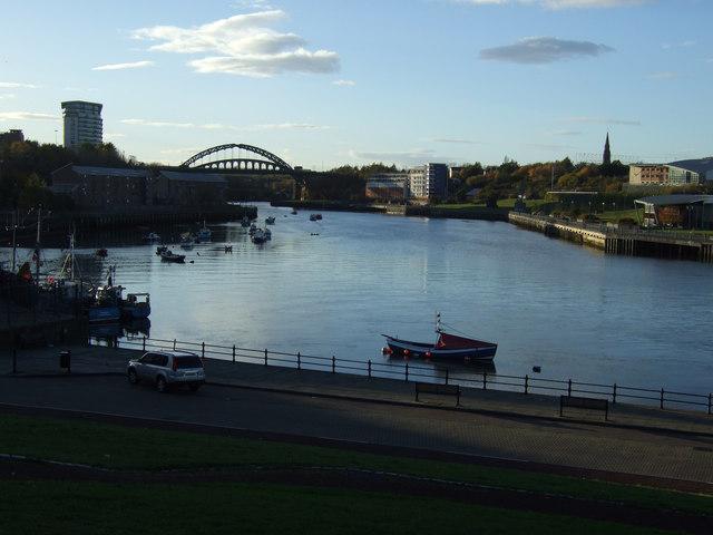 A calm river flowing through Sunderland, with a view of the Wearmouth Bridge in the distance. Small boats float in the harbor, and a red boat is docked near the riverbank. Buildings line both sides of the river, and a church spire is visible on the right. The late afternoon sunlight casts a soft glow, and the sky is mostly clear with a few clouds.