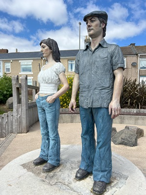 Close-up view of the small-scale replica of the "Couple" statue near the promenade at Newbiggin-by-the-Sea. The male and female figures stand side by side, dressed in casual clothing, with a residential area in the background.