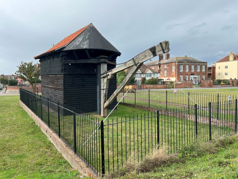 A historic wooden treadwheel crane enclosed by a black iron fence. The structure features a slanted roof and large wooden beams, set against a backdrop of houses and cloudy skies.