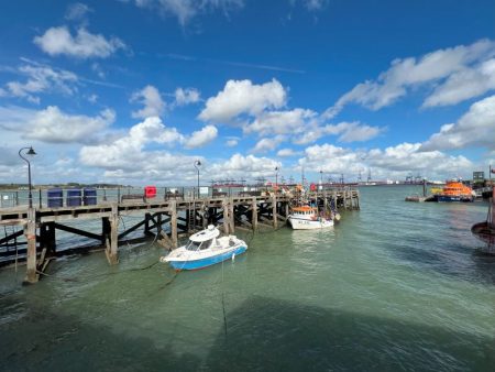 Halfpenny Pier in Harwich with moored boats under a bright blue sky, featuring a wooden dock and cranes visible in the distance