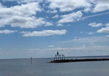 Distant view of the "Couple" sculpture by Sean Henry, located on a breakwater in Newbiggin Bay. The sculpture depicts two human figures standing close together, gazing out to sea. The sky is partly cloudy, and the calm water stretches out beneath the sculpture.