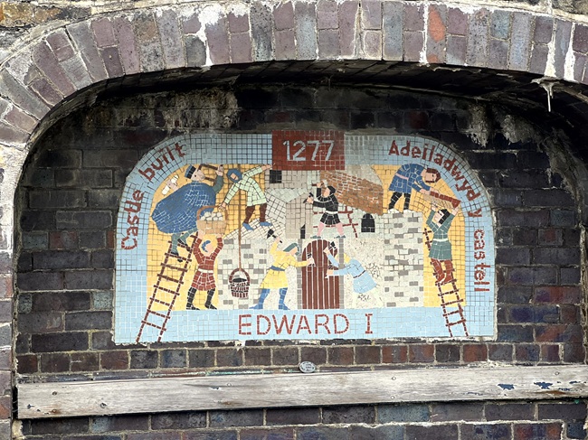 Colorful mosaic mural set in a brick arch, depicting the construction of a castle in 1277 by order of Edward I, with the text in both English and Welsh.