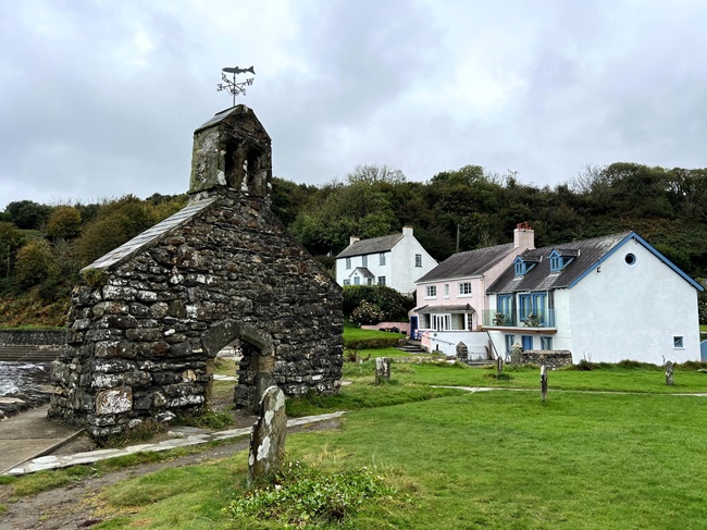 Ruins of a small stone church with a bell tower in Wales, standing near modern houses with green grass and gravestones in the foreground.