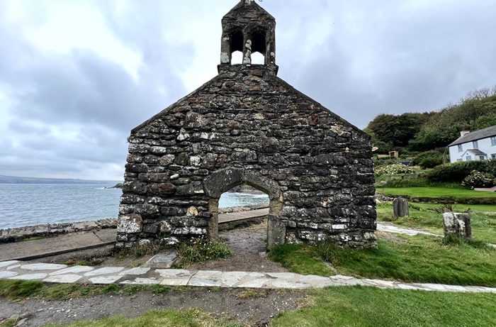Ruins of an old stone church with a bell tower, standing by the sea in Wales, with cloudy skies above and green grass around.