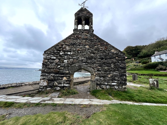 Ruins of an old stone church with a bell tower, standing by the sea in Wales, with cloudy skies above and green grass around.