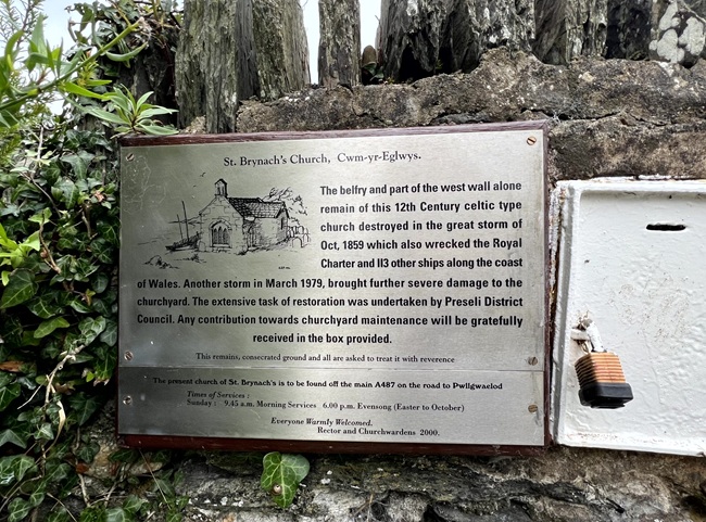 Plaque at St. Brynach’s Church, Cwm-yr-Eglwys, mounted on a stone wall with greenery around it. The plaque provides historical information about the church ruins and restoration efforts.