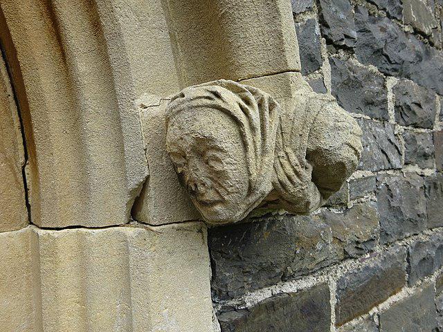 Weathered stone carving of a human face wearing a hood, protruding from a church wall.