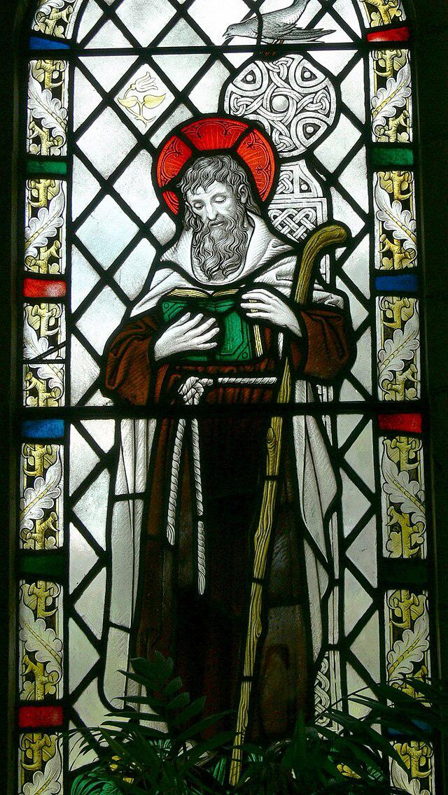 Stained glass window depicting a bearded saint in robes, holding a staff and a book, with a red halo around his head and decorative patterns in the background.
