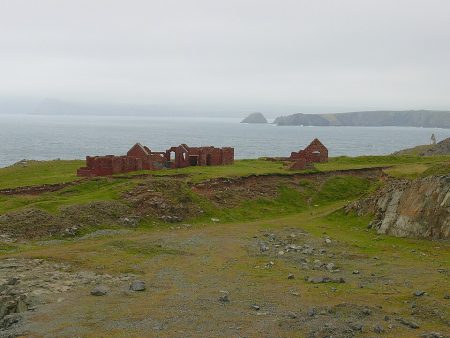 Ruins of former quarry buildings overlooking the sea at Porthgain, with red-brick walls standing on grassy ground above a rocky quarry landscape and a misty coastline in the distance.