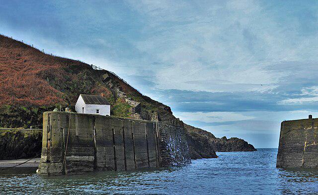 View of Porthgain Harbour showing the harbour entrance framed by high stone quay walls, a white harbour building on the left, rugged coastal cliffs, and calm sea under a cloudy sky.