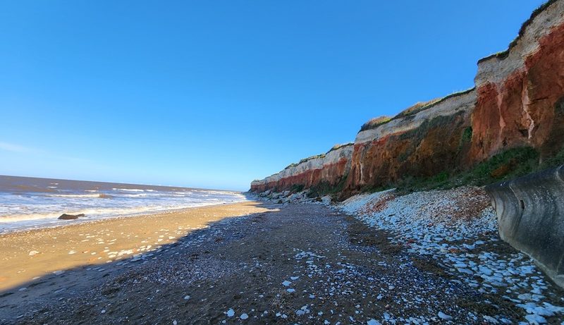 Pebble-covered beach beneath eroding red and white cliffs along a windswept coastline, with waves rolling in under a clear blue sky.