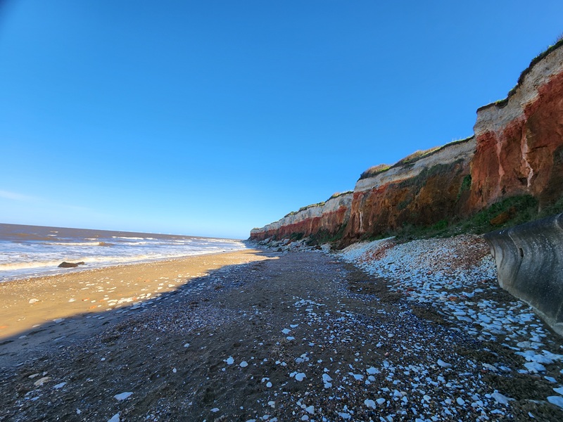 Pebble-covered beach beneath eroding red and white cliffs along a windswept coastline, with waves rolling in under a clear blue sky.