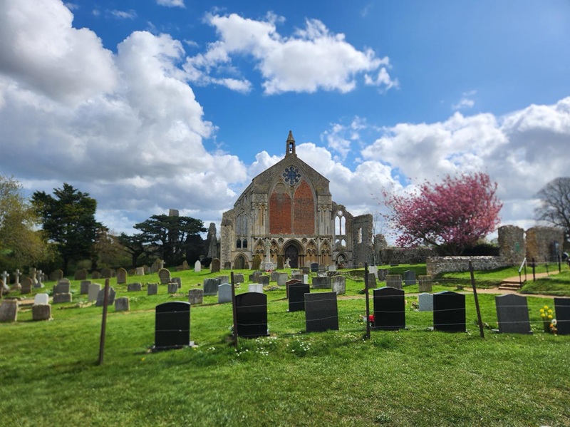 Binham Priory ruins and church facade surrounded by a historic graveyard in Norfolk, England, under a bright blue sky with dramatic clouds and blooming spring trees.
