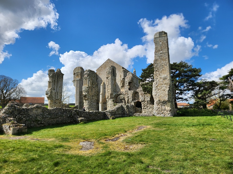 Ancient stone ruins of Binham Priory in Norfolk, England, with towering medieval walls and arches set against a bright blue sky with dramatic clouds.