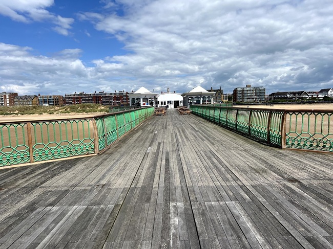 The pier at Lytham St Annes extends over the sandy beach, with distinctive green railings leading to a white pavilion under a dramatic, cloud-filled sky.