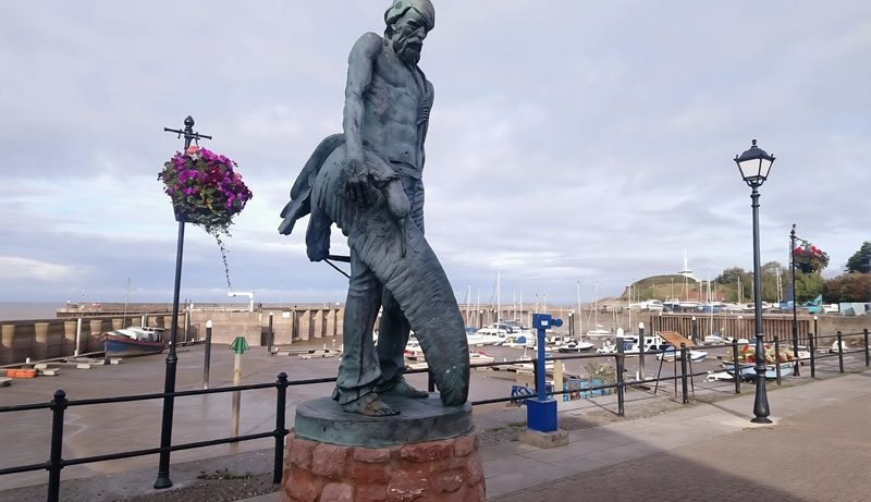 The Ancient Mariner statue overlooking Watchet Harbour in Somerset, England, with fishing boats and the marina behind.