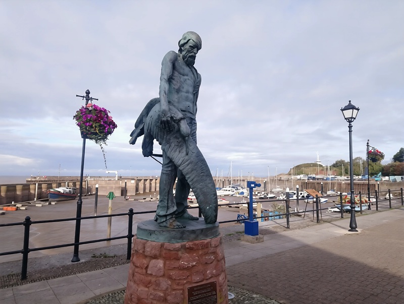 The Ancient Mariner statue overlooking Watchet Harbour in Somerset, England, with fishing boats and the marina behind.