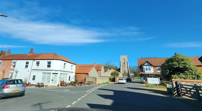 View of the village centre in Weybourne, Norfolk, with traditional cottages, a church tower, and blue skies.