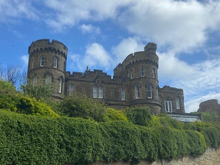 The Towers, a Victorian Gothic Revival residence in Scarborough, with castellated stone walls and round turrets surrounded by greenery beneath a partly cloudy sky, on the British coast associated with Ann Brontë.