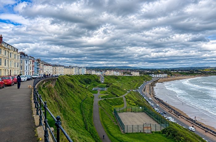 Scarborough coastline viewed from the clifftop, with colourful Victorian seaside buildings, green slopes, beach, promenade and sea beneath a dramatic cloudy sky.
