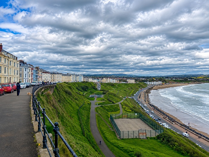 Scarborough coastline viewed from the clifftop, with colourful Victorian seaside buildings, green slopes, beach, promenade and sea beneath a dramatic cloudy sky.