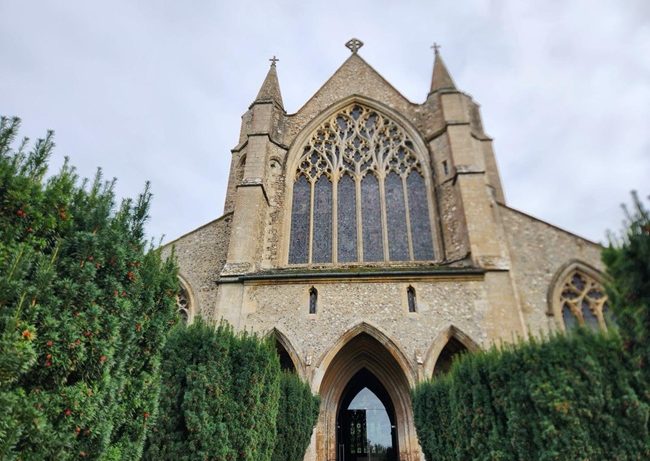 West front of St Mary's Church in Snettisham, Norfolk, showing the large Gothic window and arched entrance framed by evergreen hedges.
