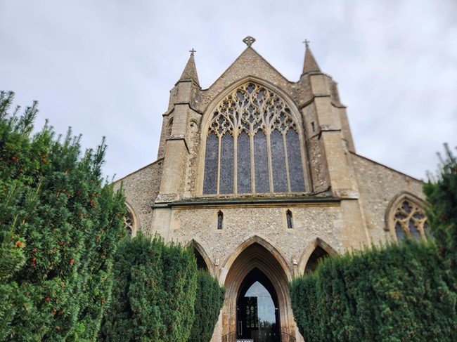 West front of St Mary's Church in Snettisham, Norfolk, showing the large Gothic window and arched entrance framed by evergreen hedges.