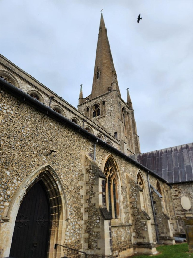 Side view of St Mary's Church in Snettisham, Norfolk, featuring the tall medieval spire and flint walls beneath an overcast sky.